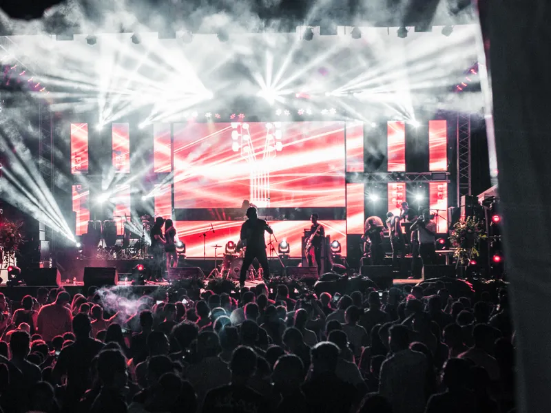 A band on a stage under white beams, red screens behind them, crowd in the foreground.