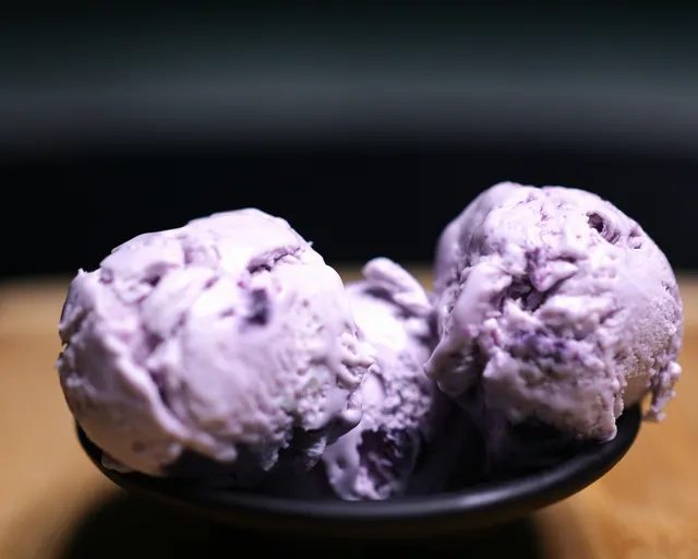 Two lilac scoops of blueberry ice cream flecked with fruit, in a dark bowl.