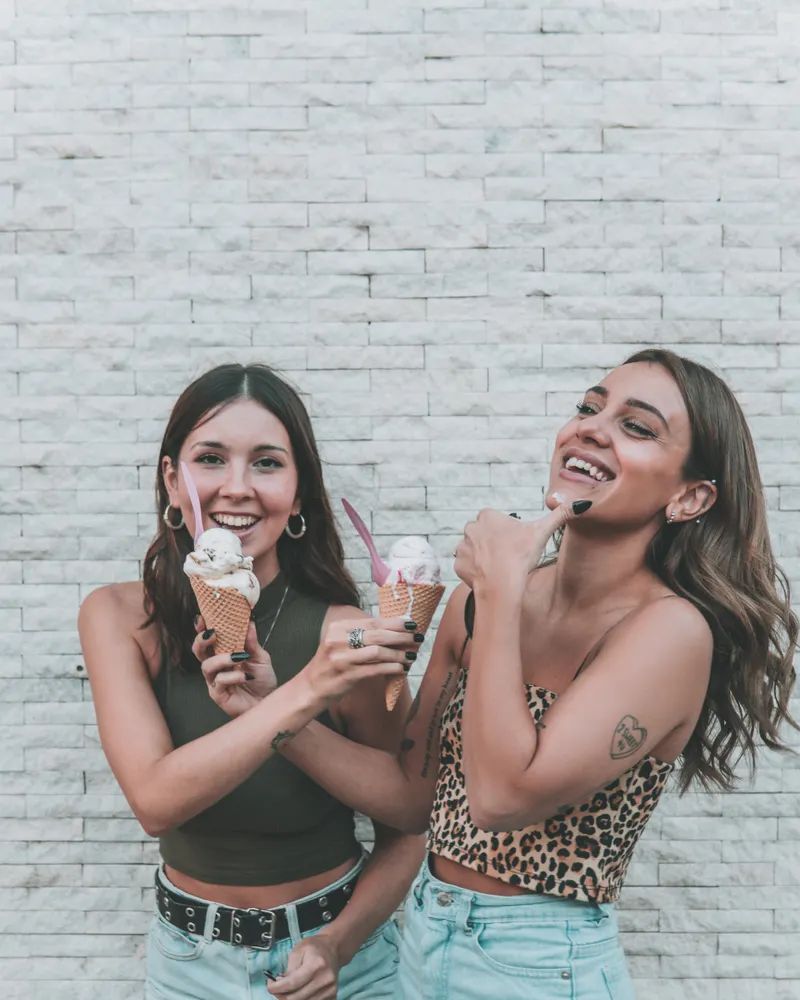 Two friends laughing against a white brick wall, each holding an ice cream cone.