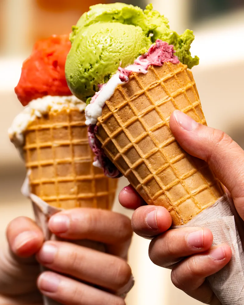 Two hands raising waffle cones, one topped with pistachio, the other with strawberry sorbet.