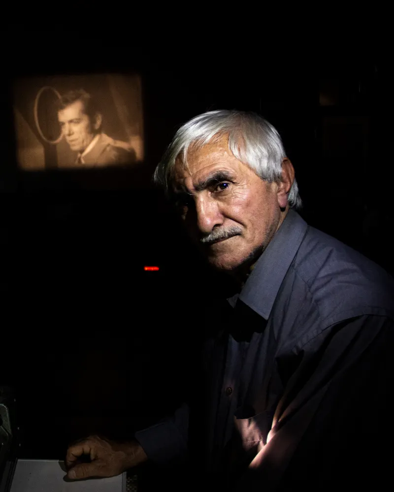 An older projectionist stands beside a vintage film projector in a dark room, a lit frame glowing on the wall behind him.