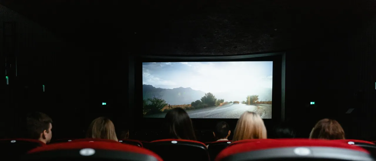 An audience in red velvet seats watches a bright screen in a darkened auditorium, seen from the back rows.