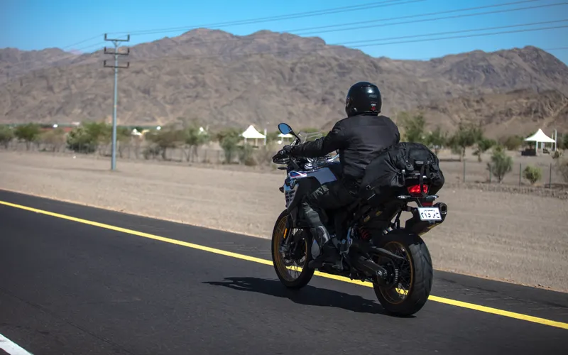 A motorcyclist rides along a sunlit road below bare brown hills.