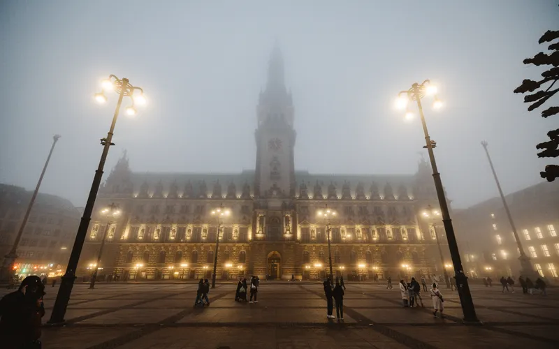 A town square at night in fog, lamps glowing, small figures crossing the paving.